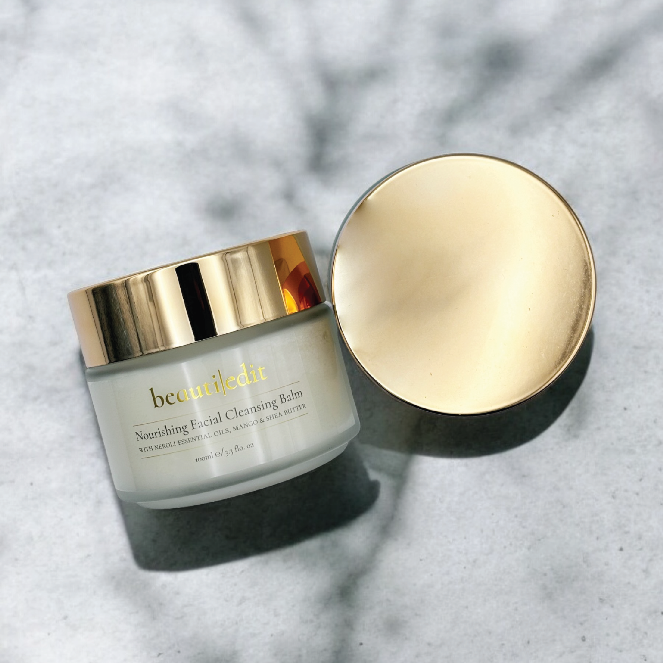 Two Cleansing balms, one showing the label and the other showing the gold top. Both on a marble counter.